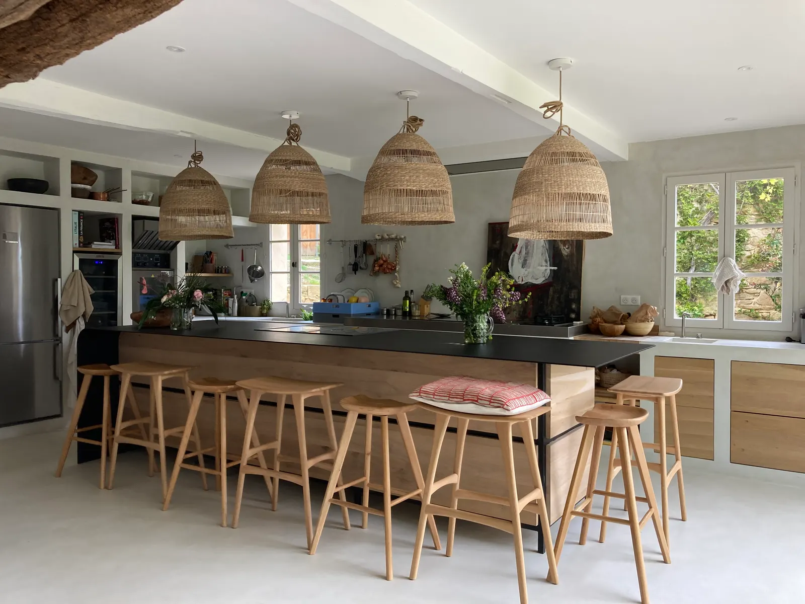 Grande cuisine ouverte avec îlot central, tabourets en bois et suspensions en fibre tressée, sur un sol en béton ciré gris clair
